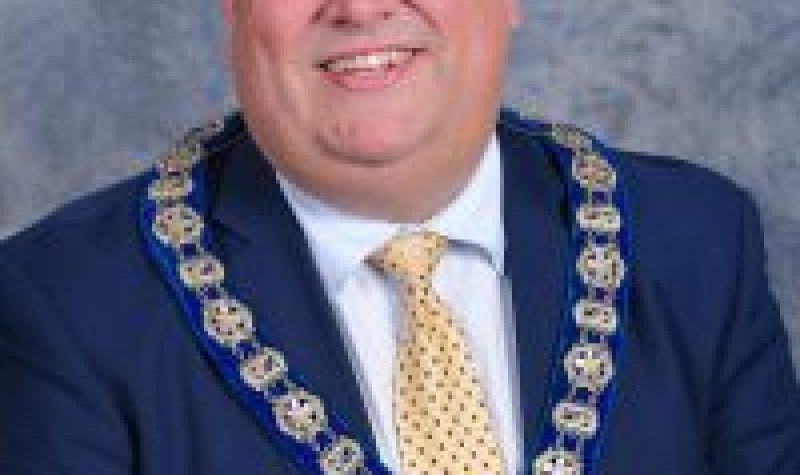 Corner Brook Mayor Jim Parsons is smiling and wearing a blue jacket and yellow tie. He is also wearing the chain of office around his neck.