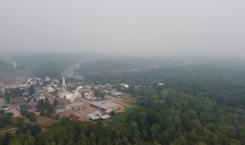 A drone photo of a small town surrounded by forest with a thick layer of smoke from forest fires.