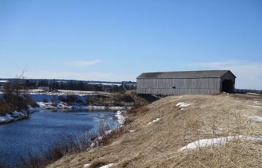 A winter landscape showing a hill and a river, and a wooden covered bridge across the river.