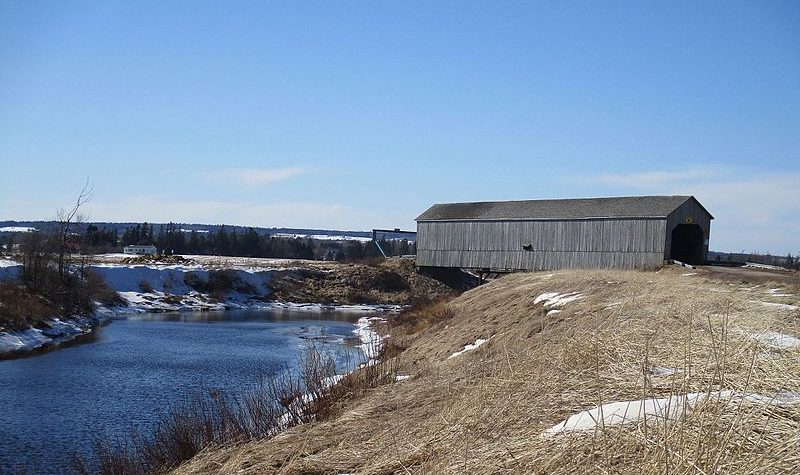 A winter landscape showing a hill and a river, and a wooden covered bridge across the river.