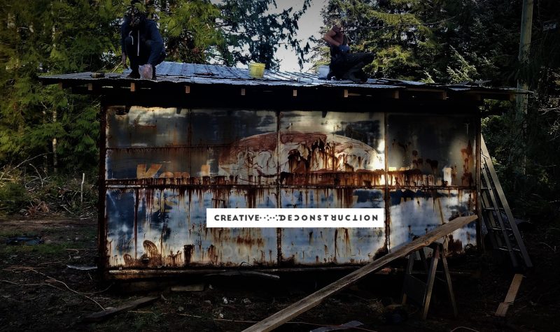 Two people kneel on top of an old shed, side of the shed reads “Creative Deconstruction”