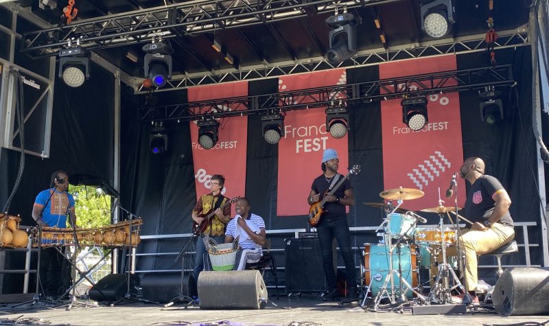 Cinq musiciens sur une scène, derrière les affichent précisent FrancoFest
