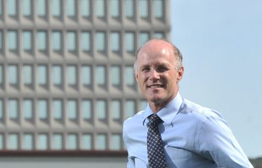 Dr Charles Gardner dehors, devant un immeuble et vêtu d'une chemise bleu pâle et d'une cravate bleu foncé