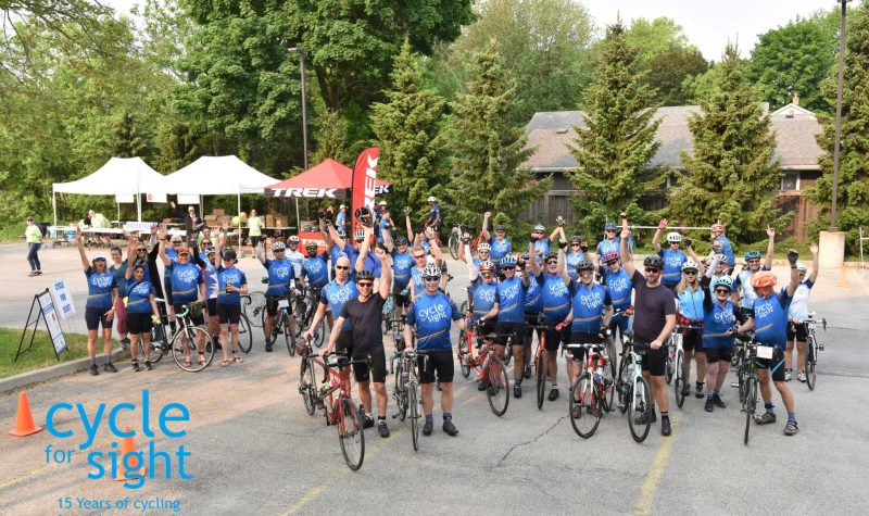 On peut voir des dizaines de cyclistes posant avec leur vélo dans un stationnement et portant le dossard bleu de l'événement « Cycle for Sight ».