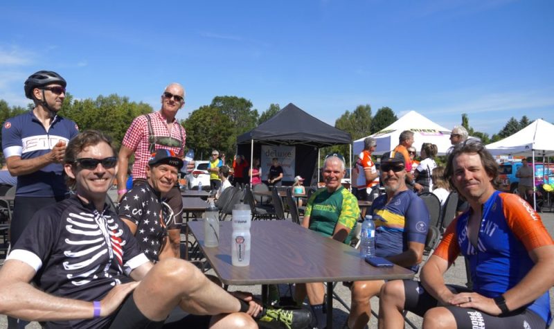 On peut voir plusieurs participants à l'événement « Cycle for Hope in Georgian Bay » de l'an dernier photographiés au Parc Rotary de Penetanguishene. Quelques participants sont souriant et assis à une table extérieure. L'ensemble des personnes portent un équipement de vélo. À l'arrière on peut voir les tentes des bénévoles.
