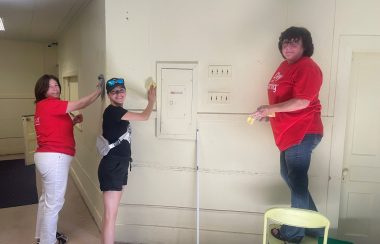 Three people, two in red, on in black scrubbing a old white wall.