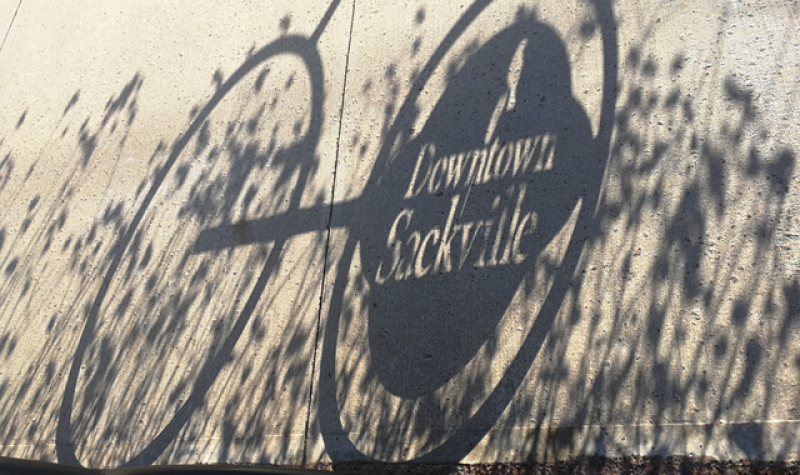 A shadow cast by a bike-shaped bike rack with the words Downtown Sackville cut into it, on a sidewalk.