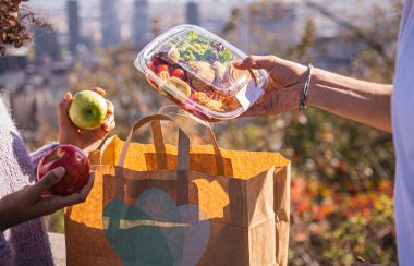 Une personne qui tient deux pommes rouge et verte, reçoit une boite remplie de tomate banane, carottes et de du poulet. Il le dépose dans un sac compostable.