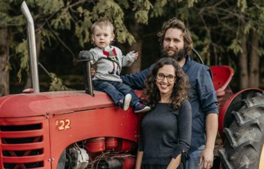 Jessica Pfisterer with husband Ryan and son Boone sit on a tractor on their family farm with woods in the back.