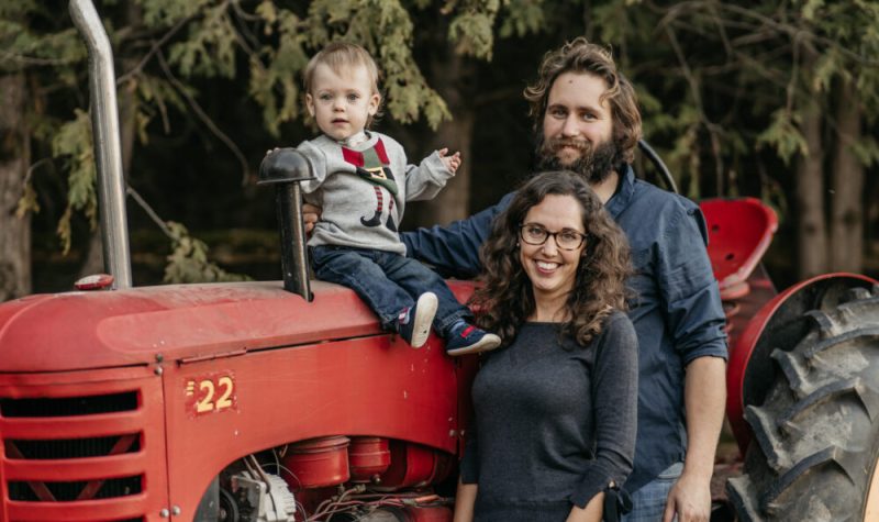 Jessica Pfisterer with husband Ryan and son Boone sit on a tractor on their family farm with woods in the back.