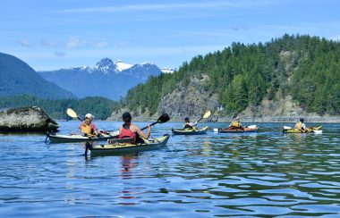 A group of people paddle kayaks on the ocean with islands and mountains in the background.