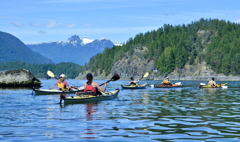 A group of people paddle kayaks on the ocean with islands and mountains in the background.