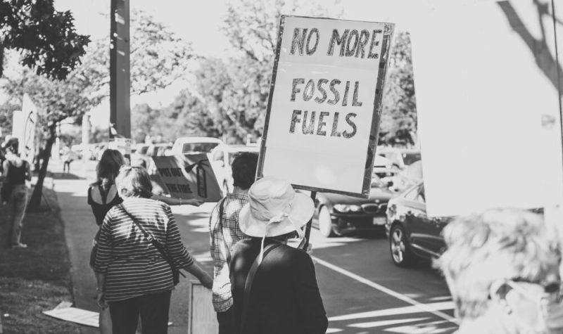Une manifestante tient une pancarte où il est inscrit « Fini les combustibles fossiles » en anglais dans une foule, devant des voitures passantes en noir et blanc.