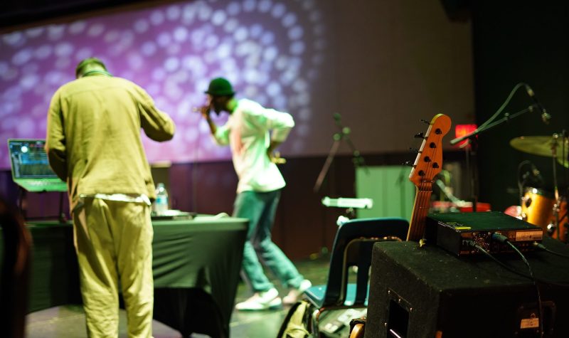Deux artistes se produisent devant une foule, avec des instruments placés derrière eux et des lumières violettes sur le mur.