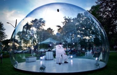 A person in all white sits in the middle of a snowglobe-like structure in the middle of a park in Kingston. The sun is setting and trees can be seen in the backdrop.