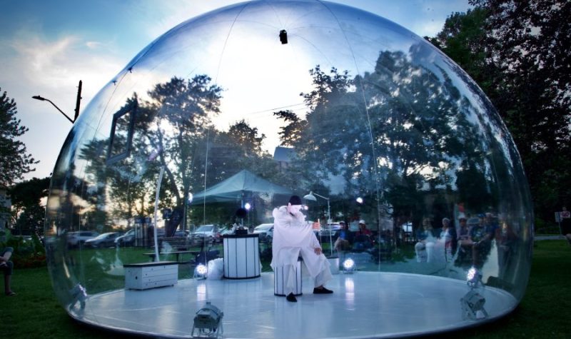 A person in all white sits in the middle of a snowglobe-like structure in the middle of a park in Kingston. The sun is setting and trees can be seen in the backdrop.