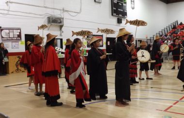 Salmon festival traditional indigenous dancers drumming and wearing regalia. They are standing in a brightly lit school gym.