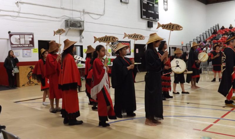 Salmon festival traditional indigenous dancers drumming and wearing regalia. They are standing in a brightly lit school gym.