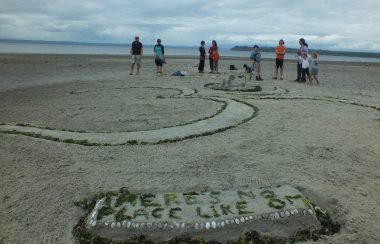 The Om symbol sculpted into the sand on a beach with a placard made of seaweed reading, “THERE”S NO PLACE LIKE OM”.