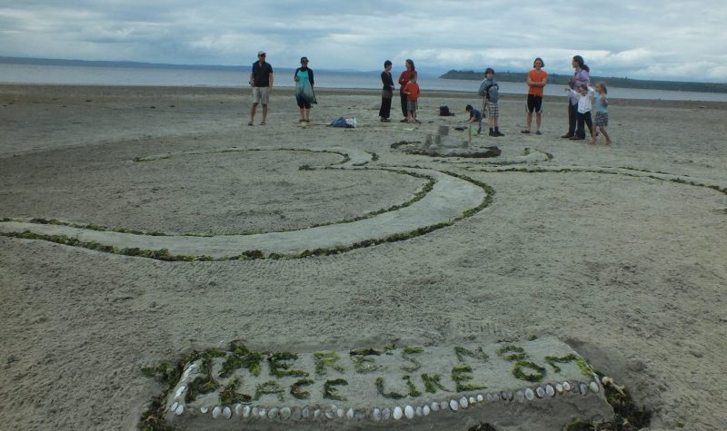 The Om symbol sculpted into the sand on a beach with a placard made of seaweed reading, “THERE”S NO PLACE LIKE OM”.