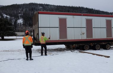 Two men wearing orange and yellow jackets help move in a trailer into a snow-covered park in Smithers on an overcast day.