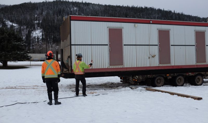 Two men wearing orange and yellow jackets help move in a trailer into a snow-covered park in Smithers on an overcast day.