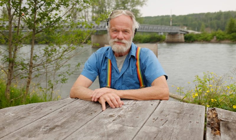 a man sits at a table outside with a bridge behind him.