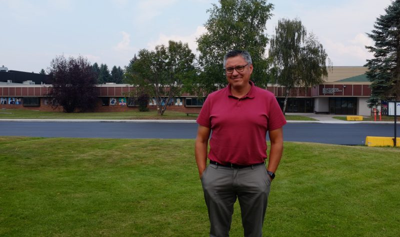A man is standing on a lawn with his hands in his pocket looking directly into the camera with a slight smile. Behind him is a one storey school building (Smithers Secondary School)