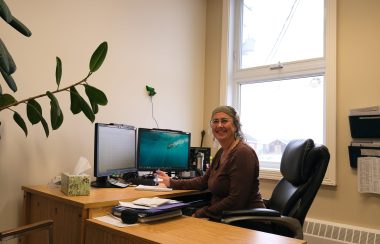 Isabelle Plante est assise à son bureau, devant son ordinateur. Les murs crèmes de son bureau sont illuminés par la fenêtre derrière elle.