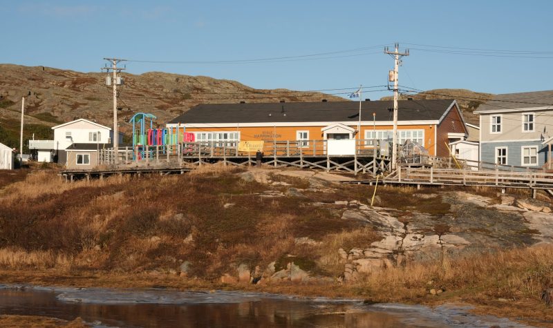 Sous un ciel bleu, le bâtiment orange de l’école de Harrington Harbour se dresse sur son morne rocheux, des structures de jeux pour enfants multicolores devant son entrée. Les trottoirs de bois caractéristiques du village sont visibles à l’avant plan