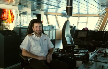 Le capitaine Corey Deveau est assis, souriant, dans la timonerie du navire, sur un siège à haut dossier et devant le tableau de bord. Par les grandes fenêtres, la lumière matinale éclaire la salle et les Vestes de flottaison individuelles orange accrochés au mur derrière lui