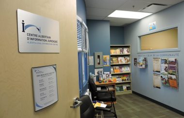 Locaux de l'Association des juristes d'expression française de l'Alberta. Des catalogues et dépliants sont oferts sur une étagère. Les murs du local sont bleu poudre et beige.
