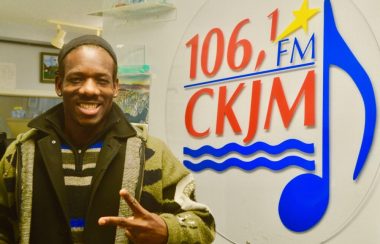 Artiste noir portant chandail brun et gilet vert en avant du logo de Radio CKJM.