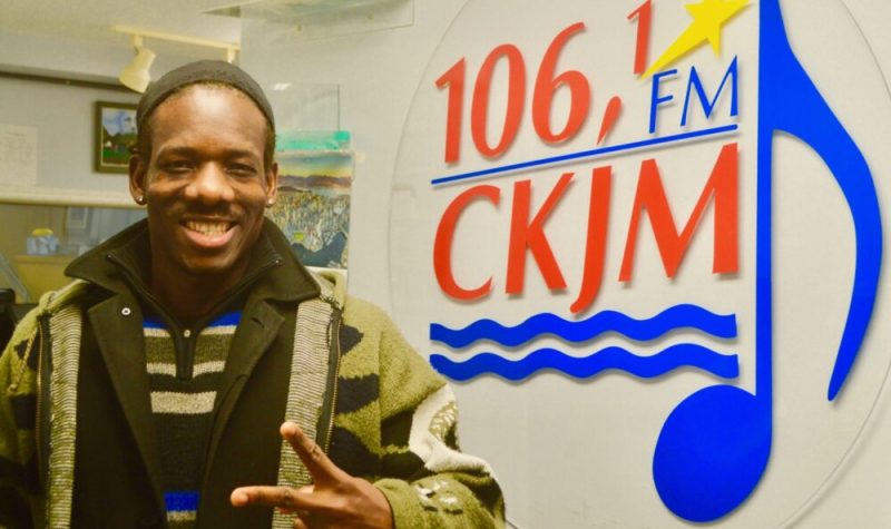 Artiste noir portant chandail brun et gilet vert en avant du logo de Radio CKJM.