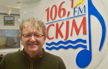 Homme avec lunettes portant un chandail noir en avant du logo de Radio CKJM.