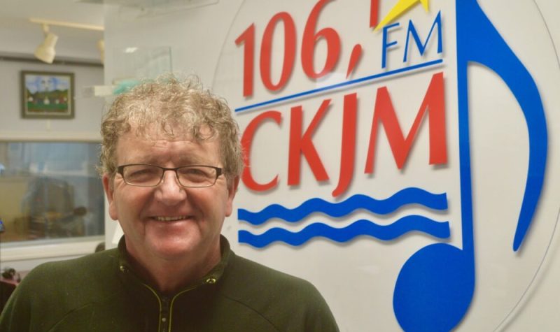 Homme avec lunettes portant un chandail noir en avant du logo de Radio CKJM.