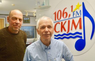 Deux hommes debout en avant du logo de Radio CKJM.