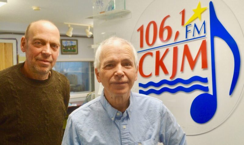 Deux hommes debout en avant du logo de Radio CKJM.