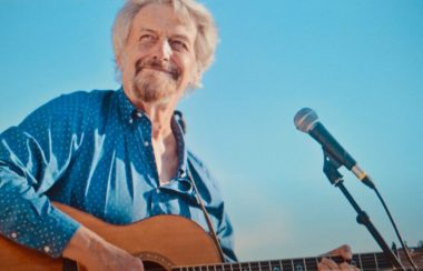 Homme avec chemise bleu jouant de la guitare et chantant dans un micro.