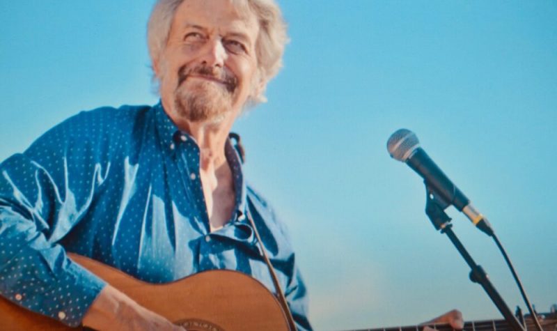 Homme avec chemise bleu jouant de la guitare et chantant dans un micro.