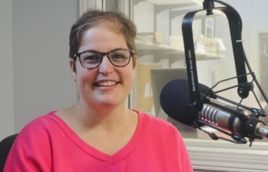 Une jeune dame avec lunettes et chandail rose en avant d'un micro dans un studio de radio.