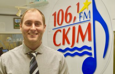 Homme avec chemise brune et cravate en avant du logo de Radio CKJM.