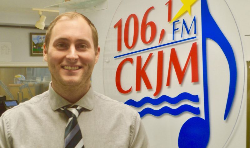 Homme avec chemise brune et cravate en avant du logo de Radio CKJM.