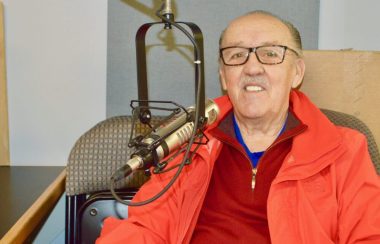 Homme avec lunettes, chandail rouge et gilet orange en arrière d'un micro de radio.