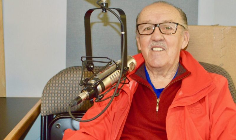 Homme avec lunettes, chandail rouge et gilet orange en arrière d'un micro de radio.