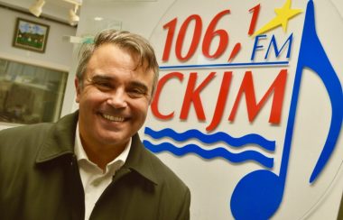 Homme souriant avec cheveux bruns, chemise blanche et gilet brun en avant du logo de Radio CKJM.