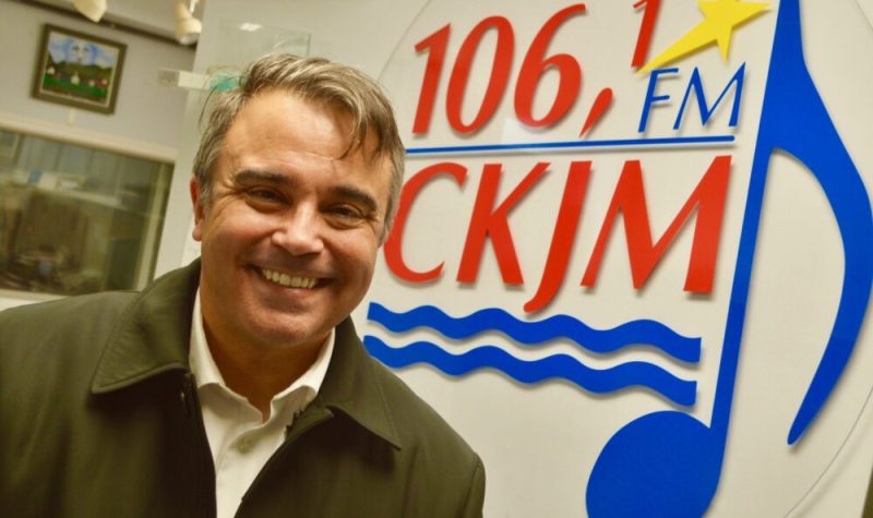 Homme souriant avec cheveux bruns, chemise blanche et gilet brun en avant du logo de Radio CKJM.