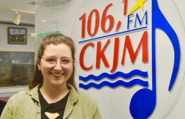 Jeune dame avec chandail noir et gilet vert en avant du logo de Radio CKJM.