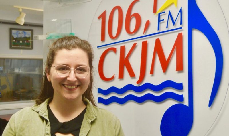 Jeune dame avec chandail noir et gilet vert en avant du logo de Radio CKJM.