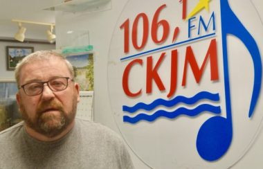 Un homme avec une barbe portant un chandail brun en avant du logo de Radio CKJM.